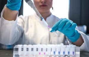 China Withheld COVID Sample From WHO, Experts Say A scientist in a lab coat examines a vial labeled with a coronavirus sample under bright laboratory lights.