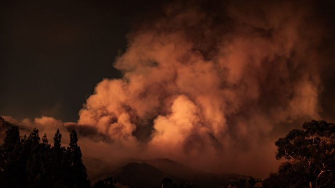 Overcrowded rescue boats packed with evacuees pull away from a burning Corfu shoreline as flames consume the island.