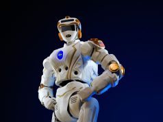 A humanoid robot running on a Beijing race course, crossing a finish line with spectators watching.