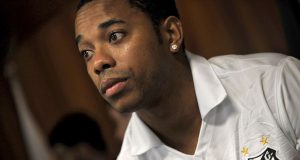 Robinho in prison uniform walks between guards inside a Brazilian jail corridor.